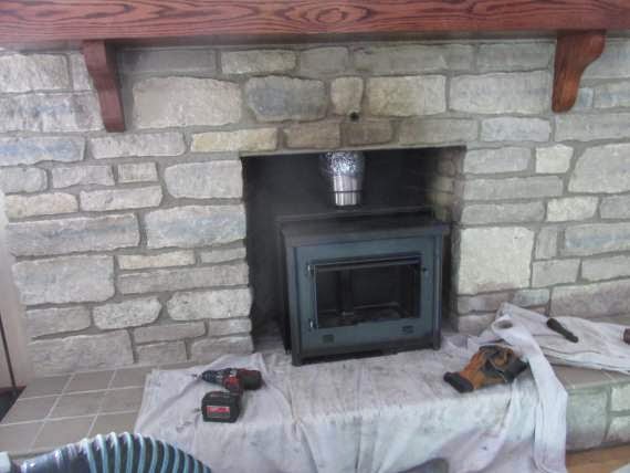 Tech in black overalls and hat working to install chimney liner in fireplace buckets surround him. It has a stone surround and a beautiful wooden mantel.