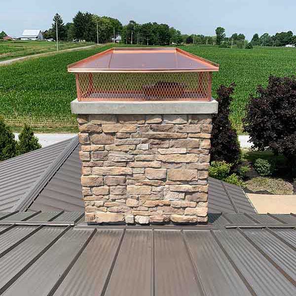 Brick chimney on home with metal chimney cover brown metal roof surrounds it and a field with trees is in the background