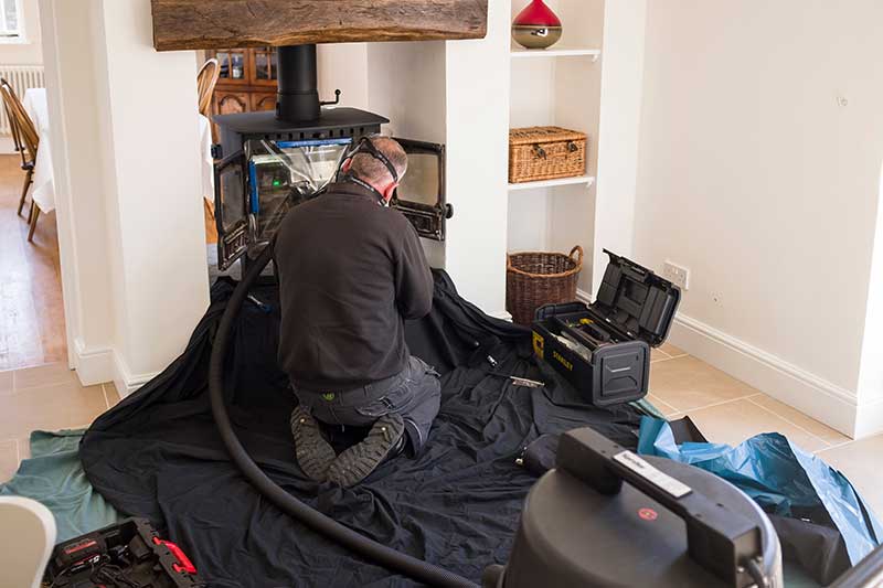 Tech on his knees sweeping wood burning stove.  He is wearing safety equipment with his tool box to his right and the vacuum to the foreground.  There are shelves to the right of the stove with baskets and a vase on the shelving.