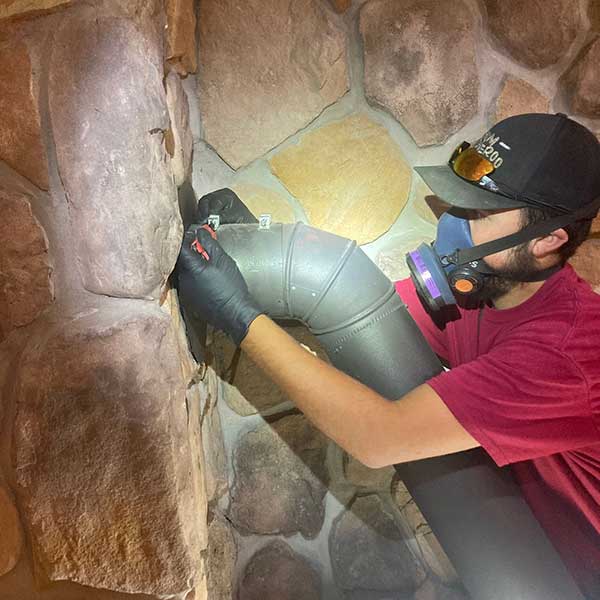 Chimney Inspections - Tech inspecting pipe running into irregular stone fireplace wearing face mask and hat.