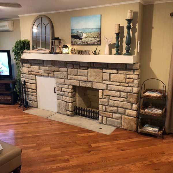 PriorFire Retrofit irregular stone fireplace with wood box and door to the left. Mantel is thick limestone and decorated with a picture, arched mirror and candles with a magazine rack to the right.